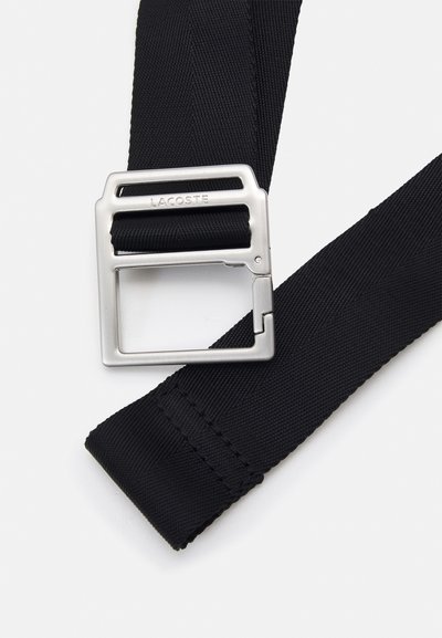 Ceinture en nylon noir dotée d'une boucle en métal argenté rectangulaire gravée "LACOSTE". La ceinture présente une finition texturée et des coutures robustes.