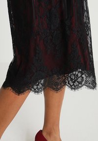 Abito in pizzo nero con uno strato interno bordeaux. Presenta motivi floreali sul pizzo e un orlo scallopato. Texture morbida con elementi trasparenti.