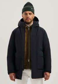 Námornická péřová bunda s kapucí, s zipovým uzávěrem vpředu, dvěma velkými náplety a hladkým povrchem, nošená přes hnědou semišovou bundu.