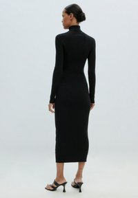 Schwarzes geripptes Midikleid mit langen Ärmeln und hohem Kragen. Enganliegende Silhouette mit geradem Saum, kombiniert mit schwarzen Absatzsandalen.