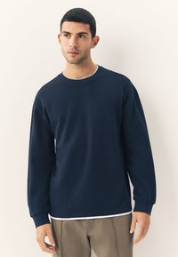 Navy long-sleeve sweatshirt in textured fabric with a crew neck and ribbed cuffs. Layered over a white hem, featuring a relaxed fit.