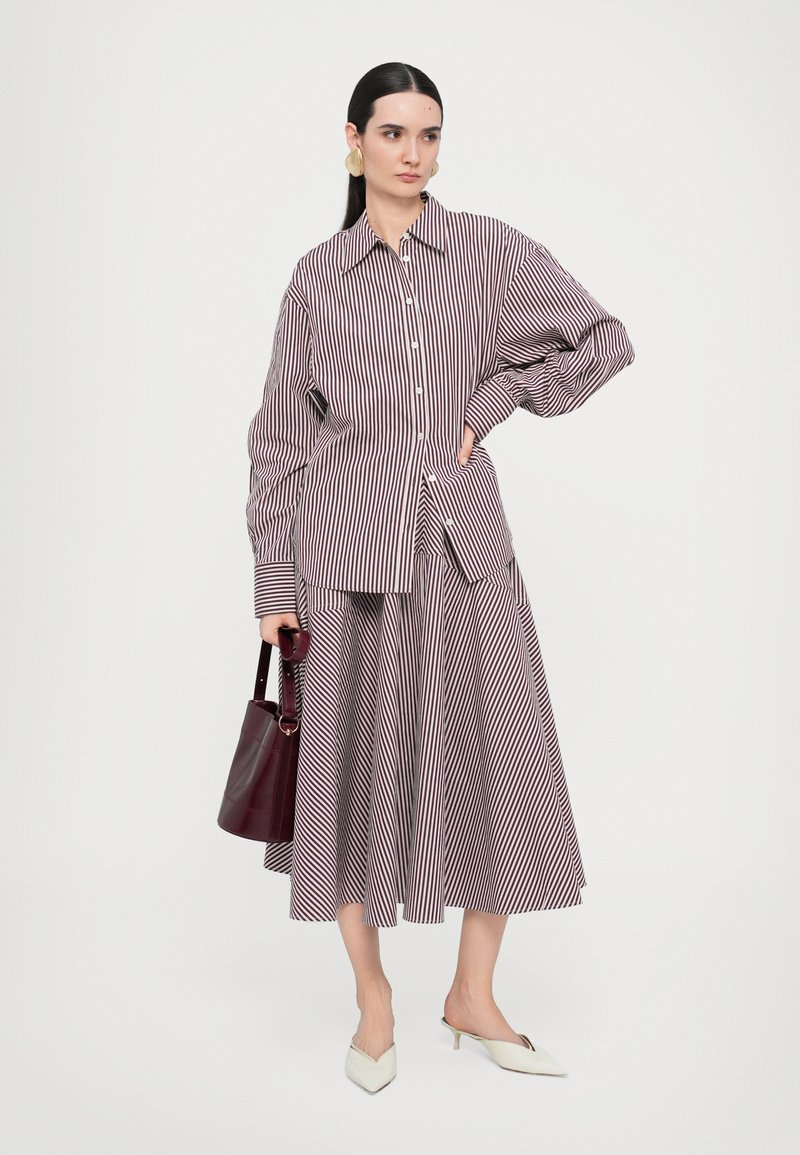 Una donna indossa una camicia a righe oversize e una gonna, tiene in mano una borsa color bordeaux e indossa tacchi bianchi, su uno sfondo bianco semplice.