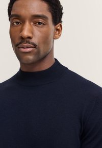 Dunkelblauer Strickpullover mit hohem, rundem Ausschnitt. Die Textur ist glatt und eng anliegend, was ein sauberes, minimalistisches Design betont.