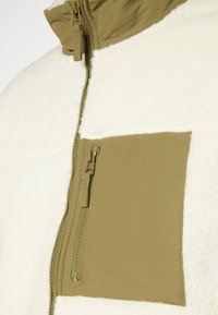 Close-up van een beige-witte fleecejas met een rits aan de voorkant en een beige ritssluiting op de borstzak, gemaakt van een zacht gestructureerde stof.