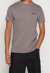 Homme portant un t-shirt gris à manches courtes avec le texte "REPLAY" sur la poitrine et un pantalon noir, une main dans la poche.