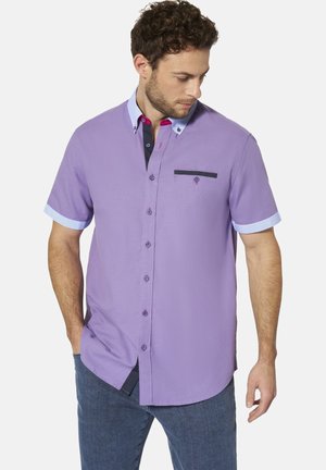 Mann trägt ein kurzärmeliges lavendelfarbenes Hemd mit hellblauen Kragen- und Manschettenakzenten, kombiniert mit blauen Jeans, schaut nach unten.