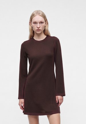 Jeune femme aux cheveux blonds raides portant une robe marron foncé à manches longues, longueur genoux, sur un fond blanc uni.