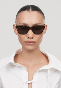 Occhiali da sole tortoise brown con forma geometrica, montatura spessa e lenti scure. Indossati con una camicia bianca a bottoni.