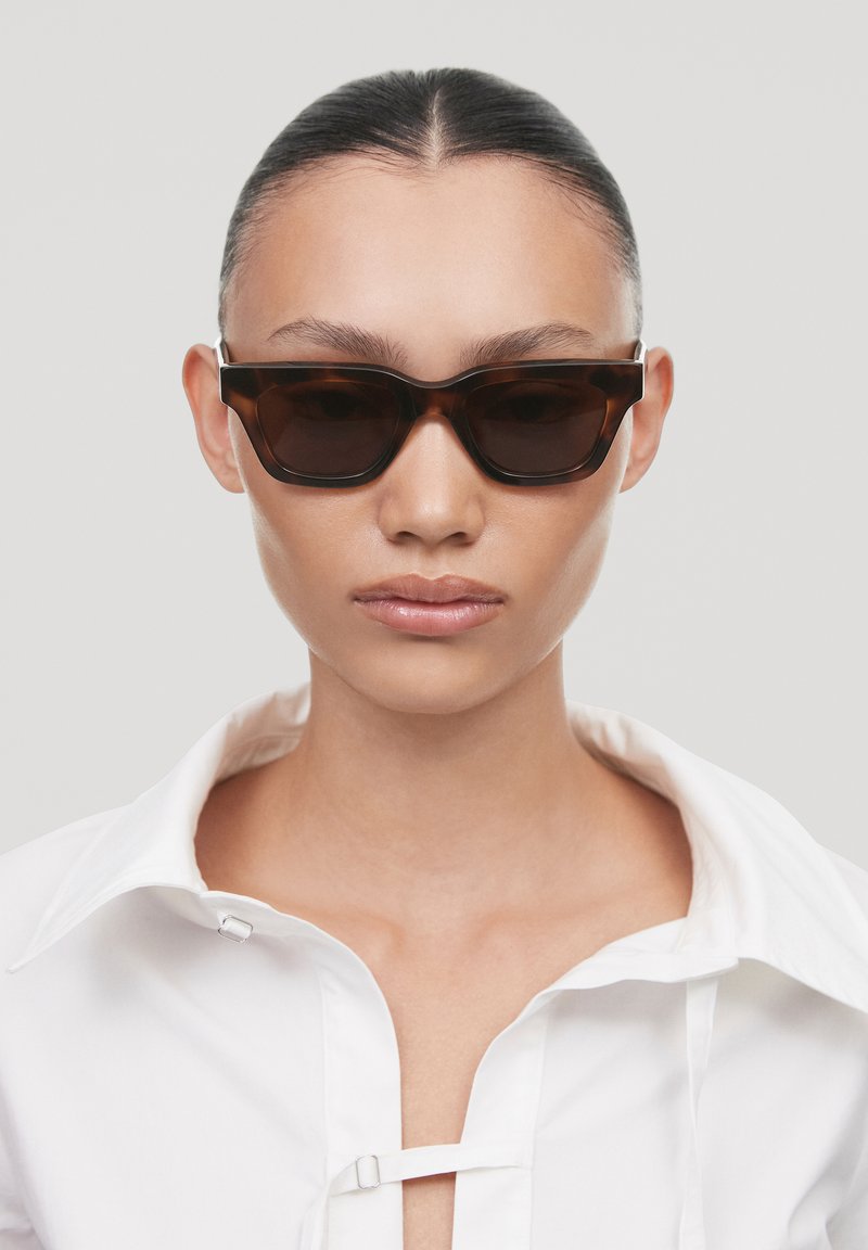 Occhiali da sole tortoise brown con forma geometrica, montatura spessa e lenti scure. Indossati con una camicia bianca a bottoni.