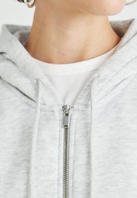 Vrat i donji dio lica osobe koja nosi srebrne kružne naušnice, bijelu majicu i svijetlo sivi zip-up hoodie s vezicama.
