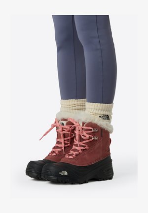 Bottes d'hiver roses en daim texturé, doublées de fourrure, avec semelles en caoutchouc noir. Portées avec des chaussettes en tricot crème et des leggings gris ajustés.