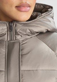 Pufferjakke i lys taupe med høj krave, synlige syninger og en lynlåslukning foran med metalhardware. Glat tekstur.