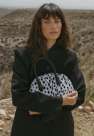 Cappotto nero, che tiene una borsa bianca con macchie nere. La borsa ha una forma curva e manici doppi con tessuto strutturato.