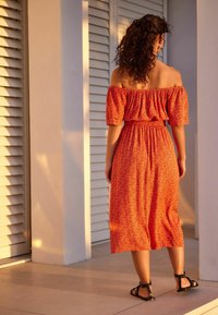 Femme aux cheveux bouclés se tient sur un sol carrelé, portant une robe midi à motifs orange décolletée et des sandales noires, faisant face à des volets blancs.