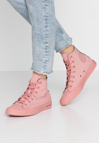 Buty sportowe typu high-top w jasnoróżowym płótnie z wzmocnionym noskiem, czarnymi akcentami i wieloma oczkami. Teksturowana gumowa podeszwa z detalem logo marki.