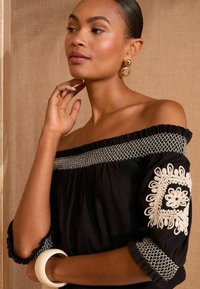Blusa nera off-the-shoulder con dettagli ricamati bianchi sulle maniche e sul collo. Tessuto morbido con bordi arricciati e accenti.