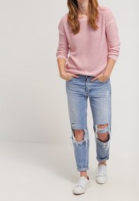 Pull rose tricoté, jean bleu clair déchiré avec de grandes déchirures aux genoux, et baskets blanches. Coupe décontractée, confortable et texture douce.