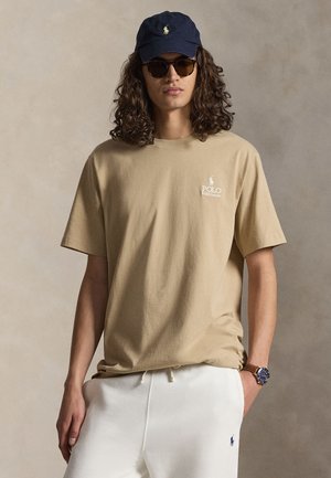 Joven con el pelo largo y rizado que lleva una camiseta polo beige, pantalones deportivos blancos, gorra azul marino, gafas de sol y reloj de pulsera, posando frente a un fondo neutro.