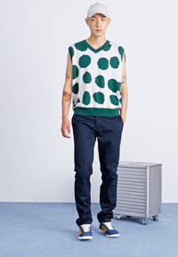 Pulover verde și alb fără mâneci, cu buline mari, purtat cu pantaloni bleumarin și adidași albaștri cu dungi galbene. Abajur deschis, fundal minimalist.