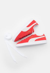 Punased sametist sneakers valgete aktsentidega, tekstureeritud kummitald ja kuldne bränding. Includes flat white laces on a light background.