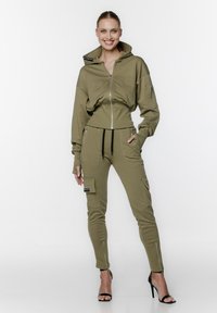 Zestaw bluzy z kapturem i spodni dresowych w kolorze oliwkowym, wykonany z miękkiego materiału. Bluza posiada wysoki kołnierz, zamek błyskawiczny oraz otwory na kciuki. Spodnie dresowe mają kieszenie cargo i zamki.