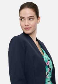 Granatowa marynarka o strukturalnym wzorze i wyłogach, w zestawieniu z zieloną sukienką w kwiaty o plisowanej teksturze i żywych kolorach.