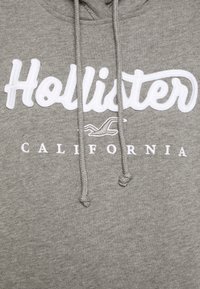 Felpa grigia in cotone con ricamo bianco della scritta "Hollister" e "CALIFORNIA" sotto. Include coulisse coordinate.