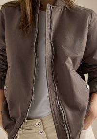 Personne portant une veste aviateur marron dézippée par-dessus une chemise blanche et un pantalon beige, les mains dans les poches, vue du cou à la taille.