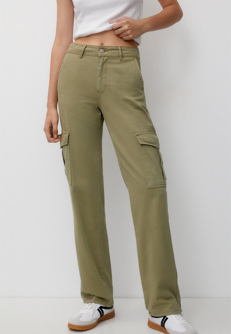 Femme portant un pantalon cargo taille haute vert olive avec poches latérales, un débardeur court blanc et des baskets blanches avec des accents noirs.