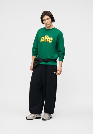 Ung person iført grøn Nike-sweatshirt, sorte vide bukser, beige Nike-sneakers og en sort bæltetaske, stående foran en ensfarvet baggrund.