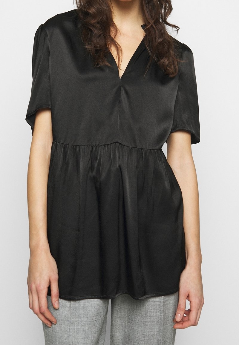 Blusa de satén negra con escote en V y mangas cortas, con una cintura fruncida y textura suave, usada sobre pantalones gris claro.