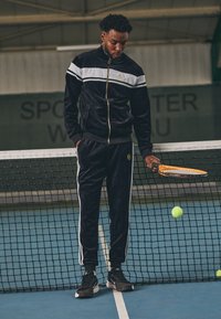 Svart velour treningsdress med hvite striper, glidelåsjakke og elastiske mansjetter. Sorte joggesko. Tenniss racket i hånden, tenniskuler i nærheten.
