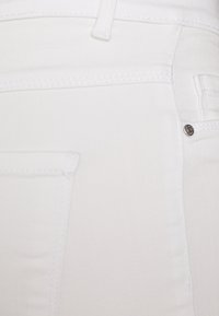 Los pantalones vaqueros de denim blanco tienen una textura suave con costuras blancas en contraste y un pequeño remache de metal plateado en el bolsillo.