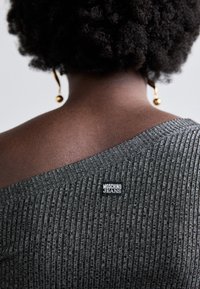 Γκρι ριγωτή μπλούζα με σχέδιο off-shoulder που διαθέτει μια μαύρη ετικέτα "MOSCHINO JEANS" πίσω, συμπληρωμένη με μια στρογγυλή σκουλαρίκι.