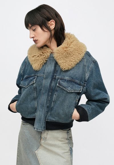 Asmuo dėvi mėlyną džinsinę striukę su dideliu smėlio spalvos apsiūtu kailiniu apykakle ir džinsinę sijoną su lopais, rankomis kišenėse.