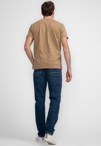 Beige bomulds t-shirt med korte ærmer, kombineret med mørkeblå denim jeans og hvide sneakers. Personen er vendt væk fra kameraet.