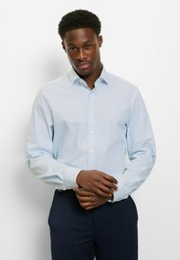 Hellblaues Hemd mit dunkelblauen Polka-Dots; hat einen spitzen Kragen und lange Ärmel, kombiniert mit dunkler Hose.