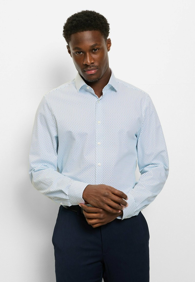 Hellblaues Hemd mit dunkelblauen Polka-Dots; hat einen spitzen Kragen und lange Ärmel, kombiniert mit dunkler Hose.