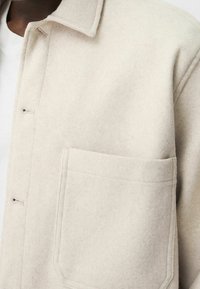 Blazer in lana beige chiaro con finitura testurizzata, dotato di colletto a punta, chiusura frontale con bottoni e una tasca sul petto.
