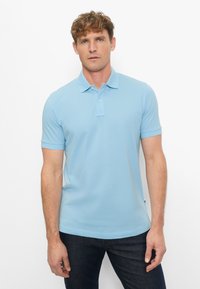 Man staat tegen een witte achtergrond, draagt een lichtblauw poloshirt met korte mouwen en donkere jeans, kijkt recht vooruit met een neutrale blik.