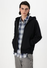 Schwarze Zip-Hoodie mit einer Kängurutasche, getragen über einem blau-weiß karierten Hemd. Das Model hat lange Haare und trägt graue strukturierte Hosen.