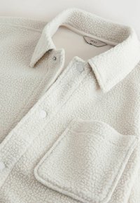 Veste-chemise en polaire blanche à surface texturée, dotée d'une fermeture à boutons sur le devant, d'une poche poitrine et d'un col classique.