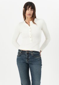 Long-sleeve, white ribbed top with a crew neckline and button closure down the front. Paired with dark blue fitted jeans.