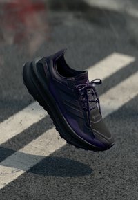 Chaussure de course violet foncé avec lacets en suspension au-dessus d'une route en asphalte avec des lignes blanches.