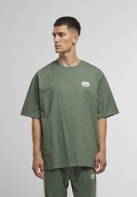 Grøn bomuld t-shirt med korte ærmer, rund hals og et lille hvidt logo på venstre bryst. Glat tekstur, afslappet pasform.