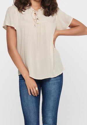 Frau trägt eine cremefarbene kurzärmelige Bluse mit Schluppe am Ausschnitt und blaue Jeans, steht mit einer Hand in der Hüfte und die andere entspannt.