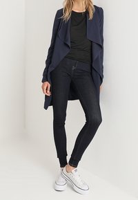 Cardigan ouvert bleu marine avec un design drapé, associé à un haut noir ajusté et un jean skinny en denim foncé, complété par des baskets blanches.