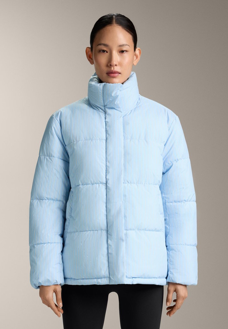 Femme portant une doudoune rayée bleu clair avec un col montant et un pantalon noir, debout devant un fond neutre.