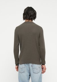 Olive green ribbed long-sleeve sweater with a crew neck, featuring knit texture and a fitted bottom hem. Worn over light blue denim jeans.