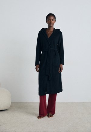 Zwarte badjas met capuchon, een riem en een voorkeurszak, gemaakt van zacht materiaal; gecombineerd met losse rode broek, staande op een licht vloerkleed.